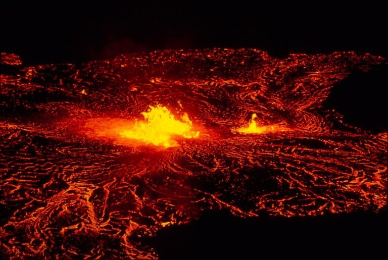 “Volcán en plena erupción expulsando columnas de fuego y lava incandescente que desciende por la ladera, iluminando el cielo oscuro con un resplandor rojizo.”